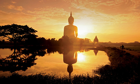 A giant buddha statue faces a temple in the distance over its reflection in still water at sunrise